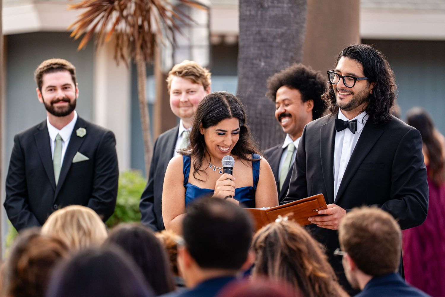 Wedding ceremony with bride, groom, and officiant smiling outdoors, groomsmen standing behind in formal attire.