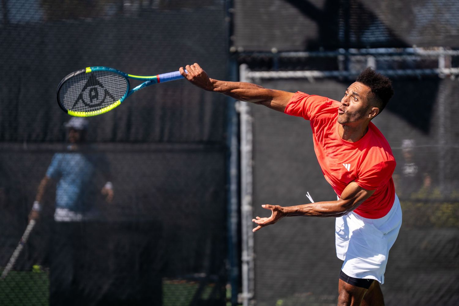 Tennis player in red shirt reaches up to serve with a Dunlop racket on an outdoor court.