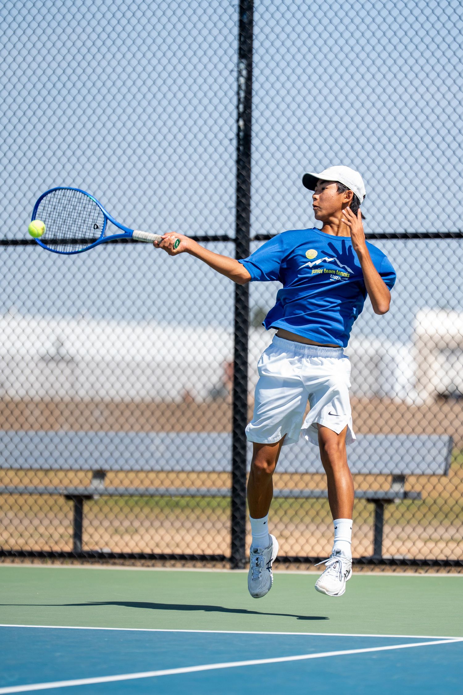 Young male tennis player in blue shirt jumps to hit a forehand on an outdoor hard court on a sunny day.