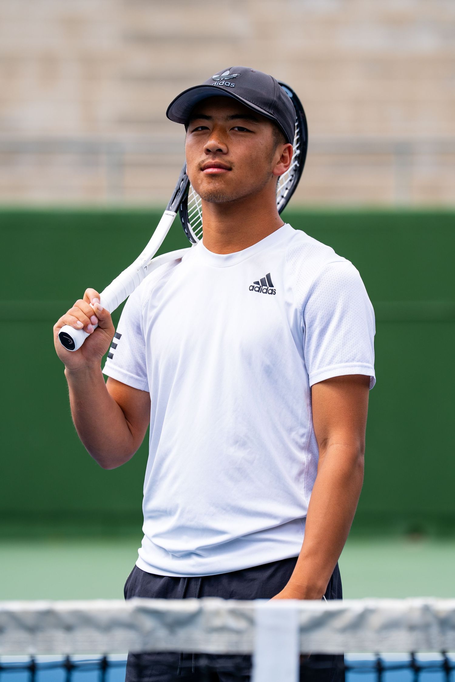 Young male tennis player in white Adidas shirt holding racket on outdoor court.