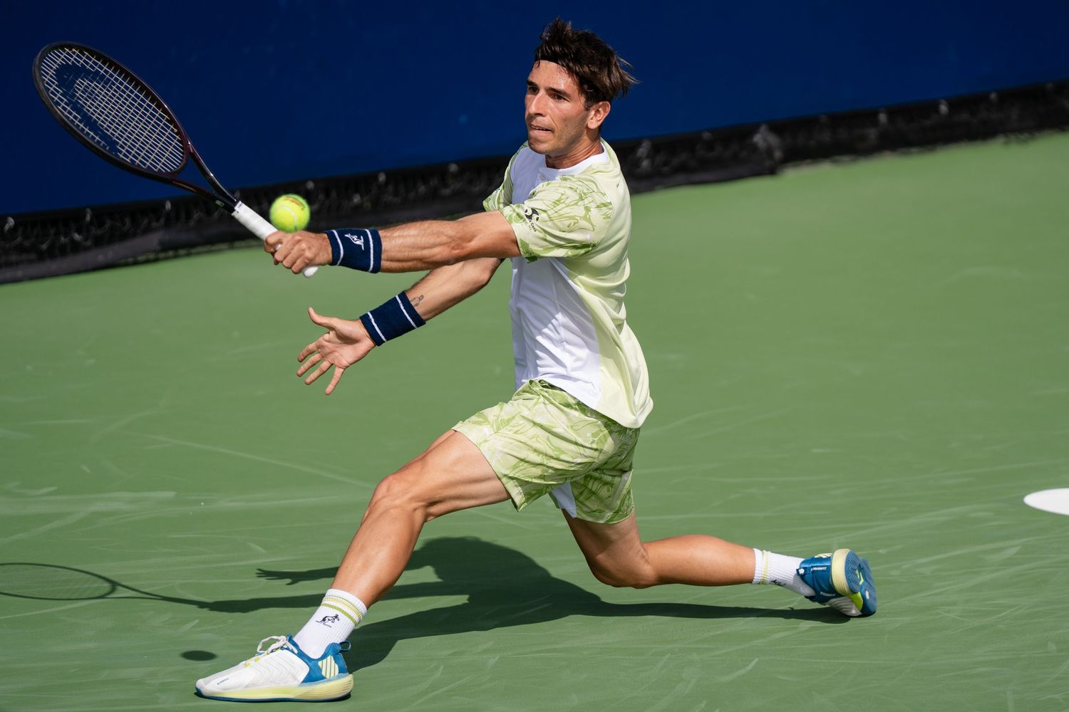 Tennis player in yellow outfit lunges for backhand shot on hard court.
