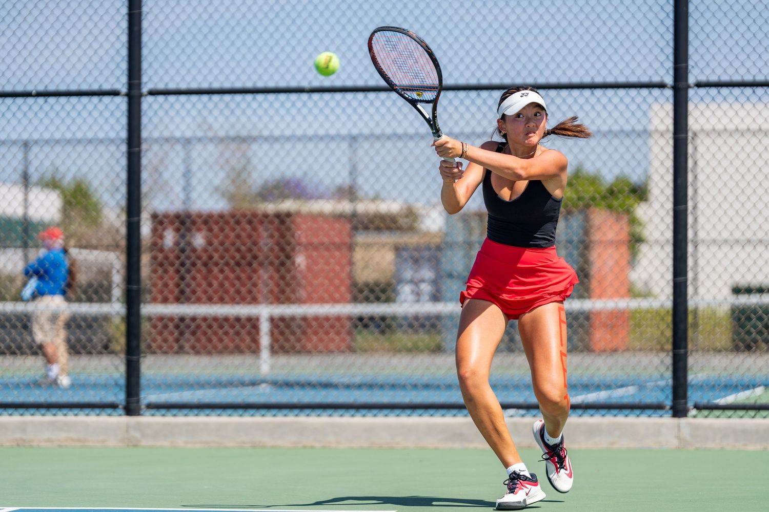 Female tennis player in red skirt executes a powerful backhand shot on an outdoor court.