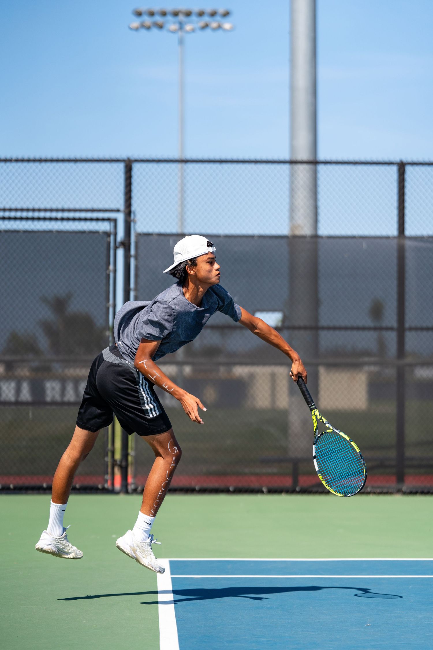 Athletic male tennis player leaps while serving on an outdoor hard court wearing gray shirt and black shorts.