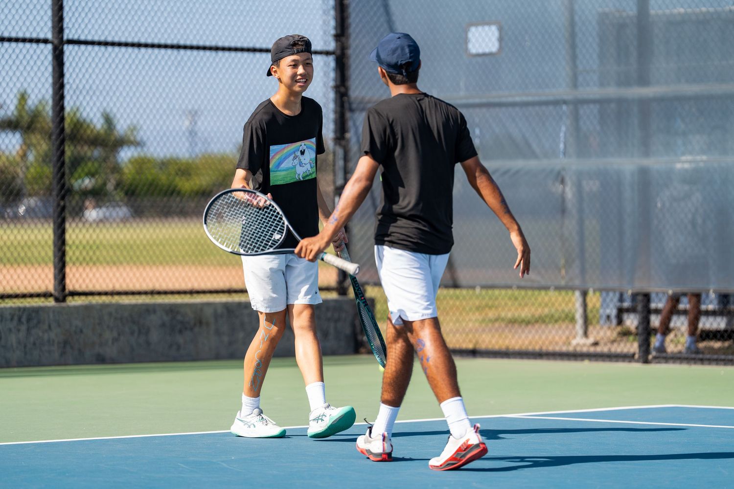 Two male tennis players in black outfits sharing a friendly handshake on an outdoor tennis court.