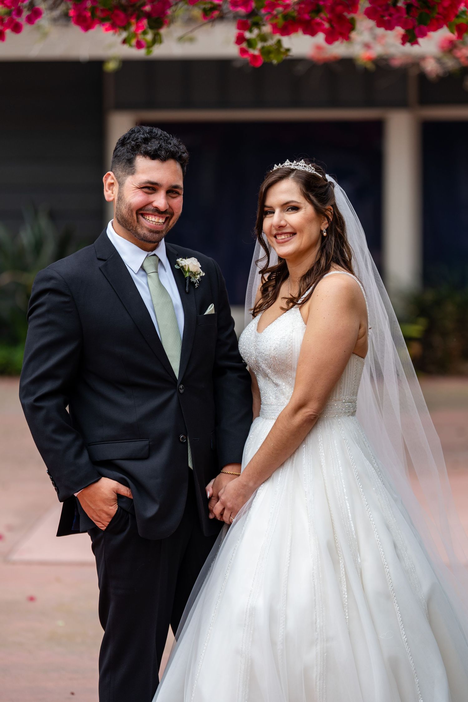 A smiling bride in a white ballgown and veil holds hands with a groom in a dark suit at an outdoor wedding.