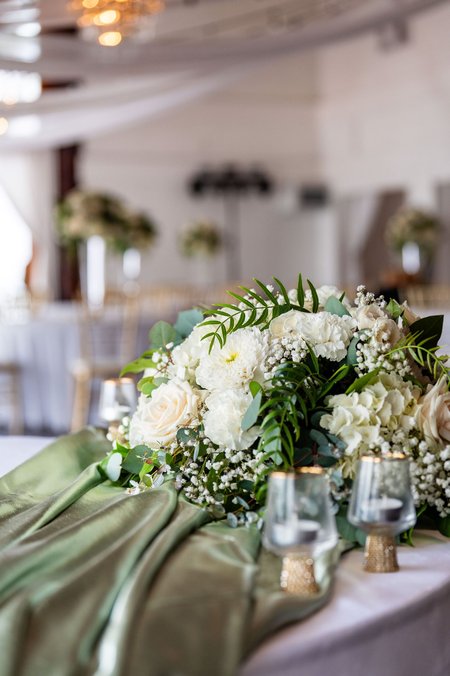 Elegant wedding reception table with white roses, hydrangeas, greenery centerpiece, sage green runner, and glass candle holders.