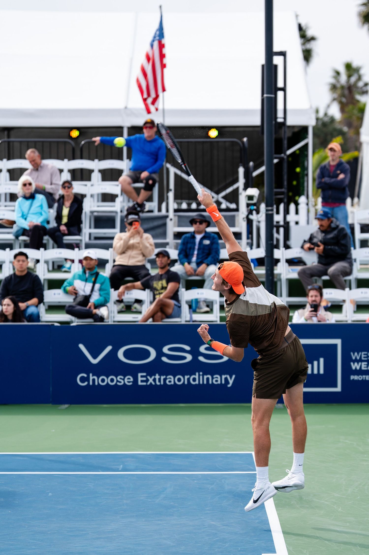 Male tennis player in brown outfit serves ball at VOSS San Diego Open tennis tournament.