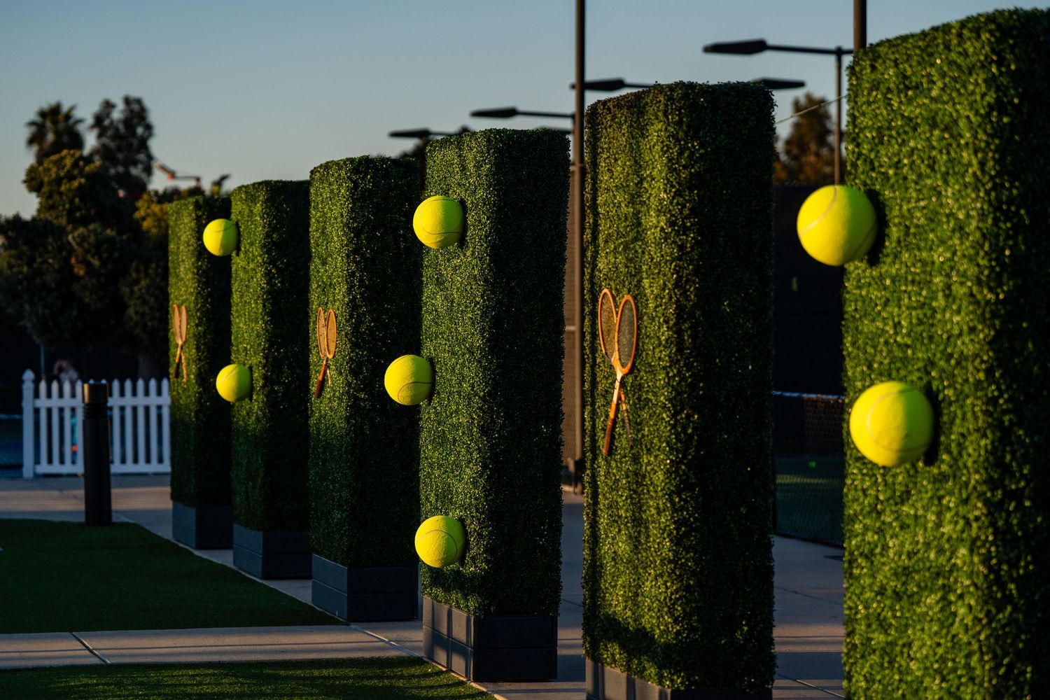 Decorative hedge columns with oversized tennis balls and rackets line a pathway at a tennis venue.