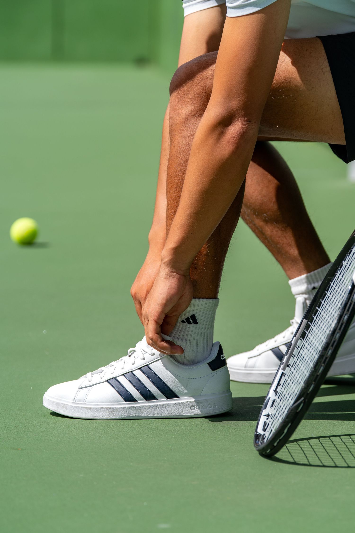 Tennis player tying white Adidas sneakers on a green hard court with a tennis ball nearby.