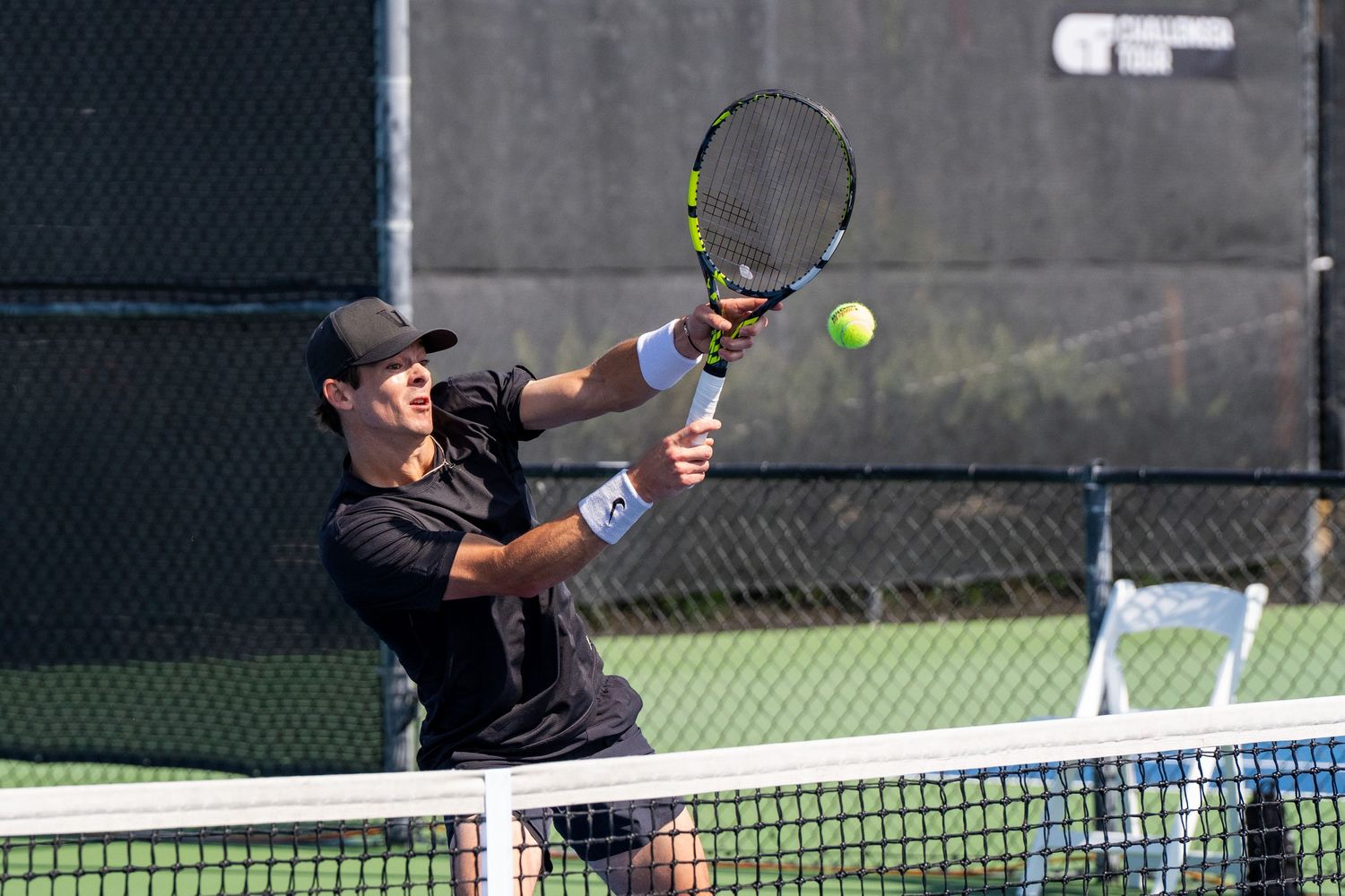 Male tennis player in black hits an overhead smash near the net on an outdoor court.