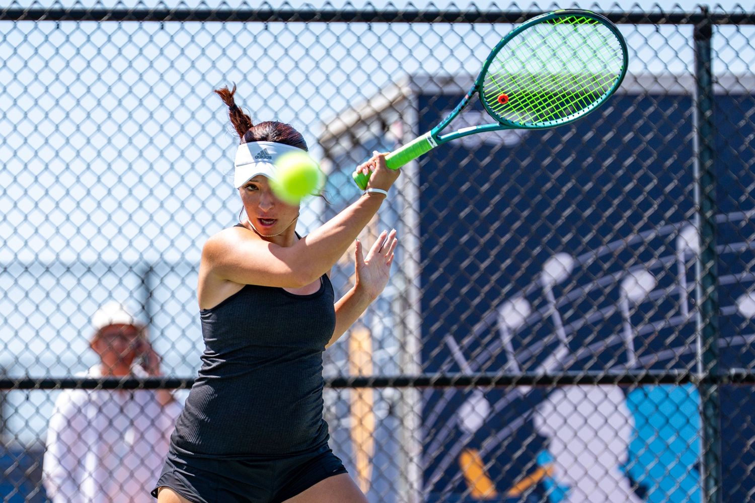 Female tennis player in black outfit strikes a forehand volley on a sunny outdoor court.