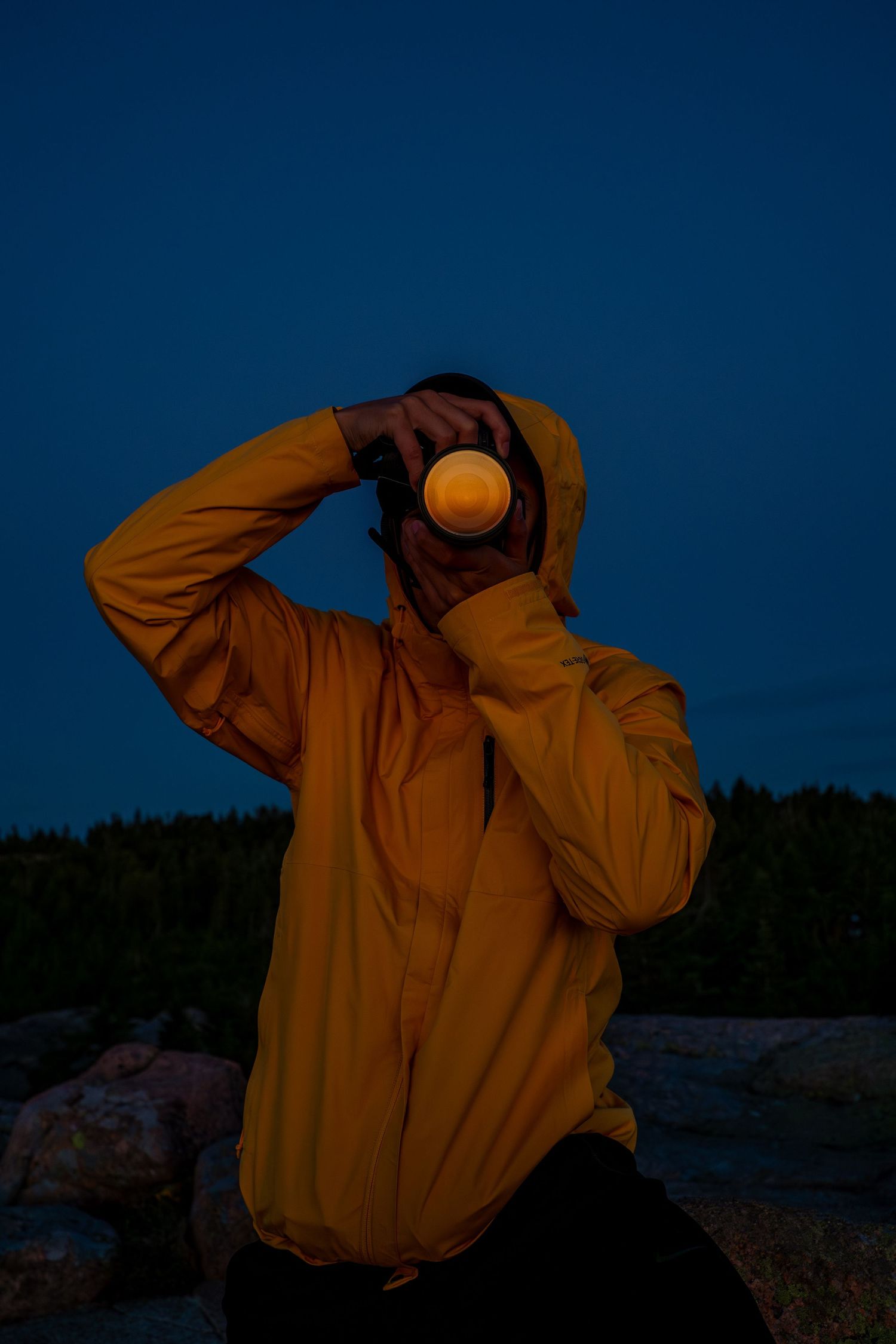 Person in yellow hooded jacket photographing at night with camera lens glowing against dark blue sky.