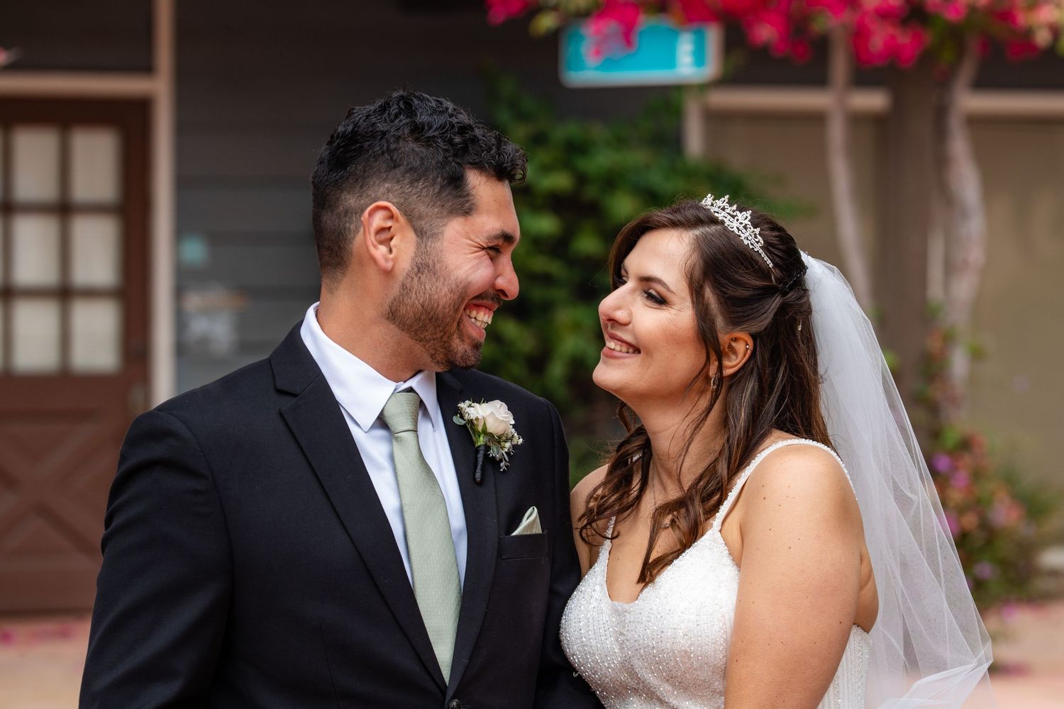 A smiling bride and groom share a joyful moment outdoors, with the bride wearing a veil and tiara on their wedding day.