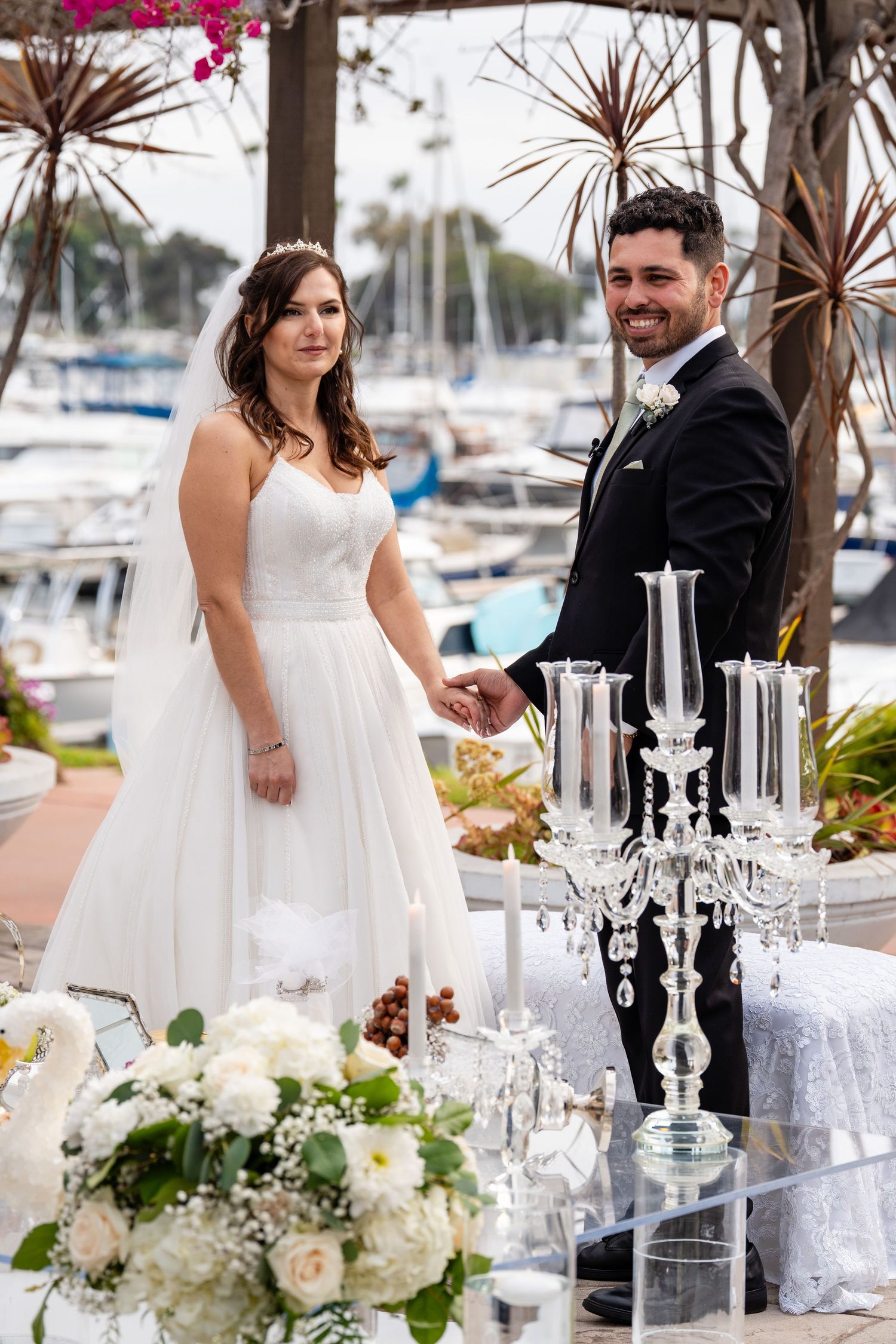 Bride and groom holding hands at outdoor marina wedding ceremony with white floral arrangements and crystal candelabra.