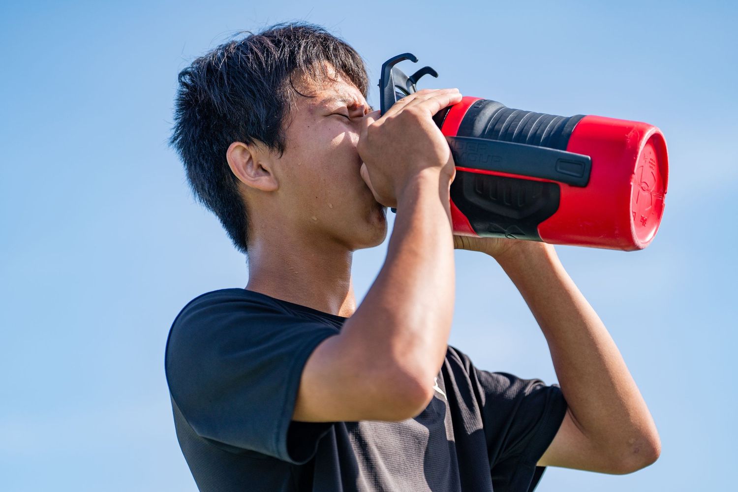 Young male athlete drinking from a large red water bottle against a blue sky.