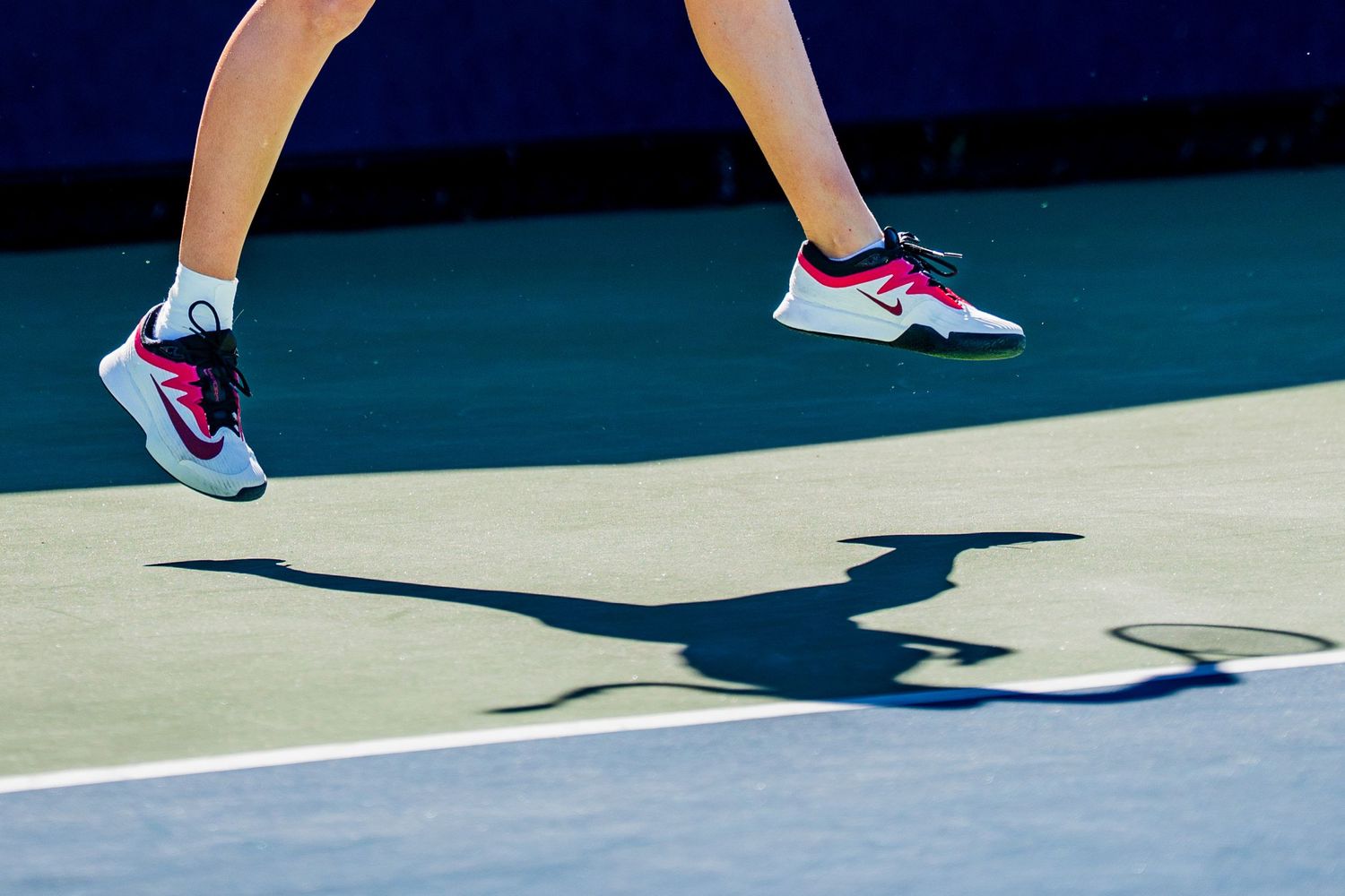 Close-up of a female tennis player jumping in pink and white Nike shoes on a blue tennis court.