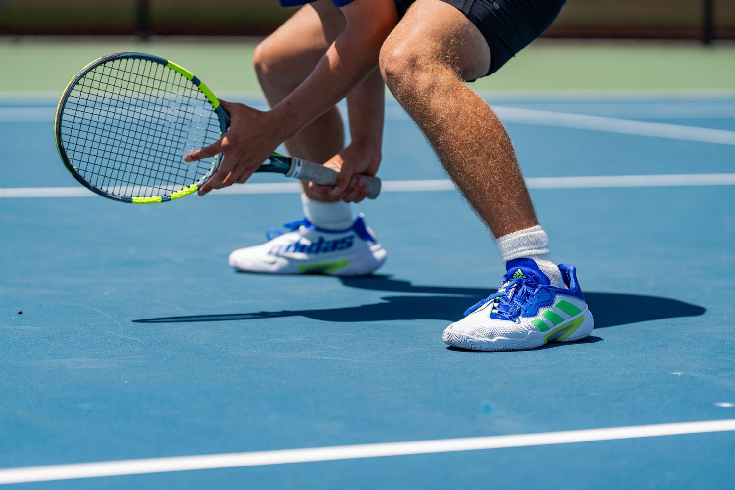 Close-up of a tennis player crouching low with a racket on a bright blue outdoor court.