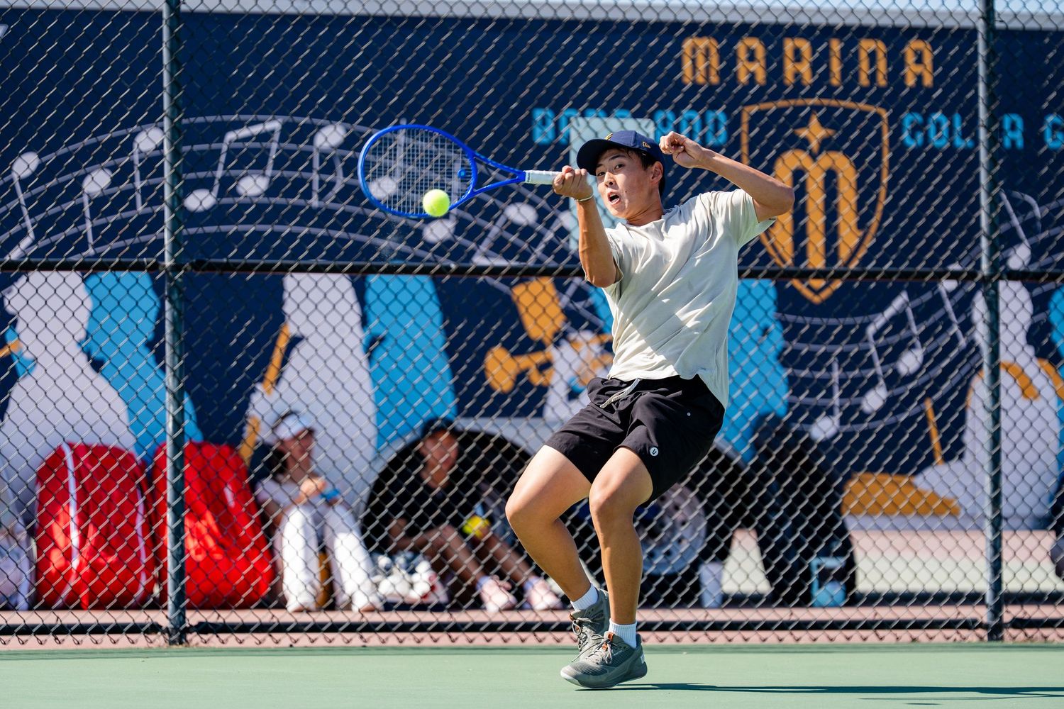 Young tennis player hits a forehand in front of a Marina High School banner on a bright sunny day.