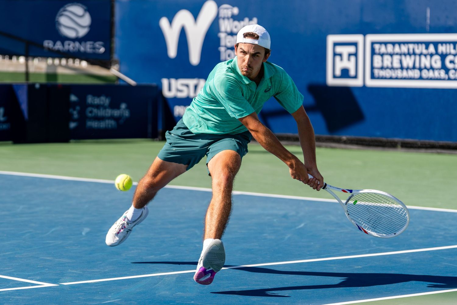 Male tennis player in teal shirt lunging to hit a low backhand on a blue court.
