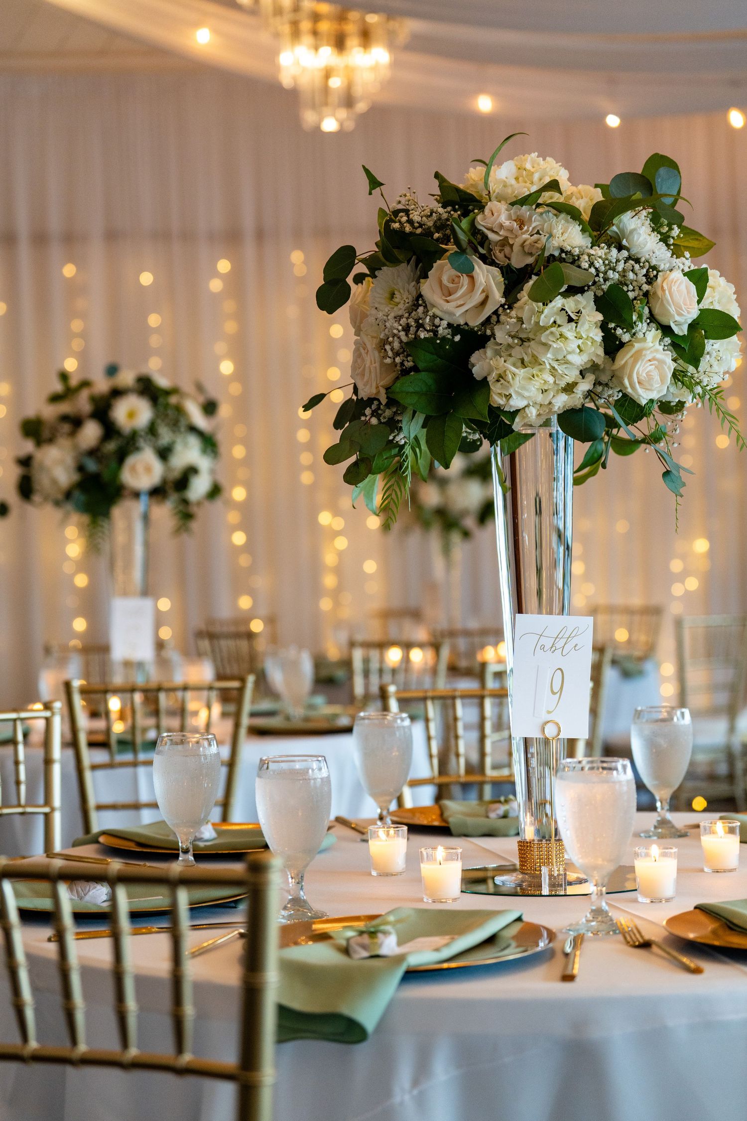 Elegant wedding reception table with tall floral centerpiece of white roses, hydrangeas, and greenery, gold chairs, and candlelight.