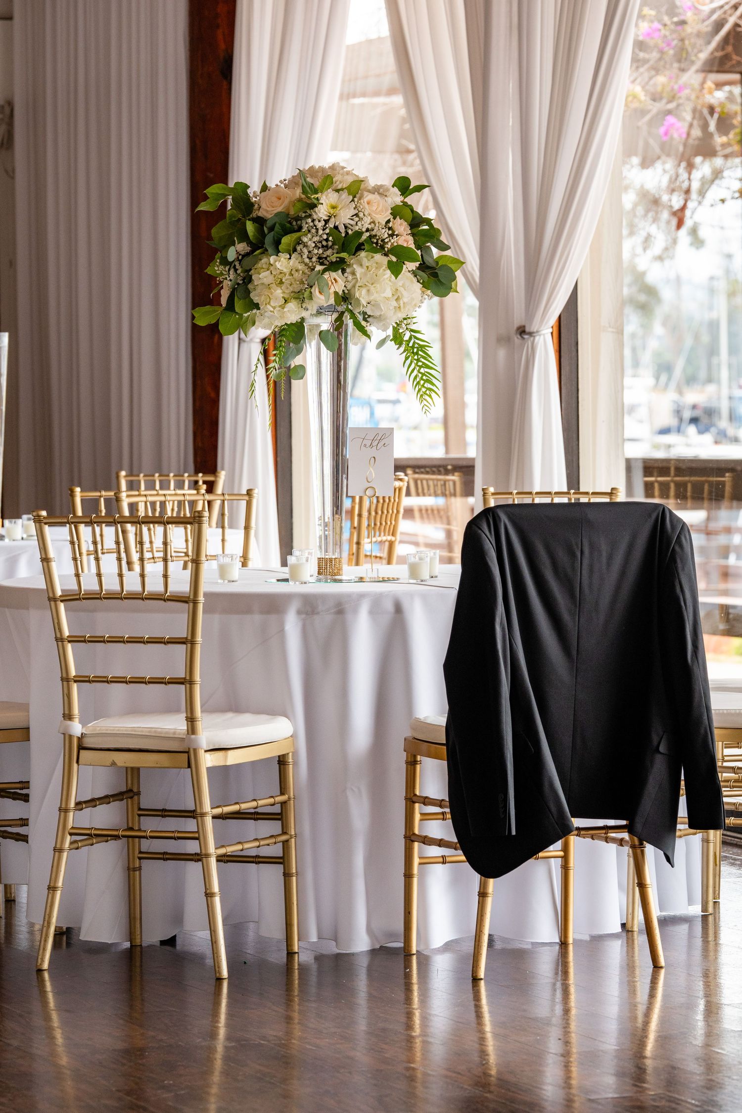 Elegant wedding reception table with gold chiavari chairs, white linens, and a tall floral centerpiece in a draped venue.