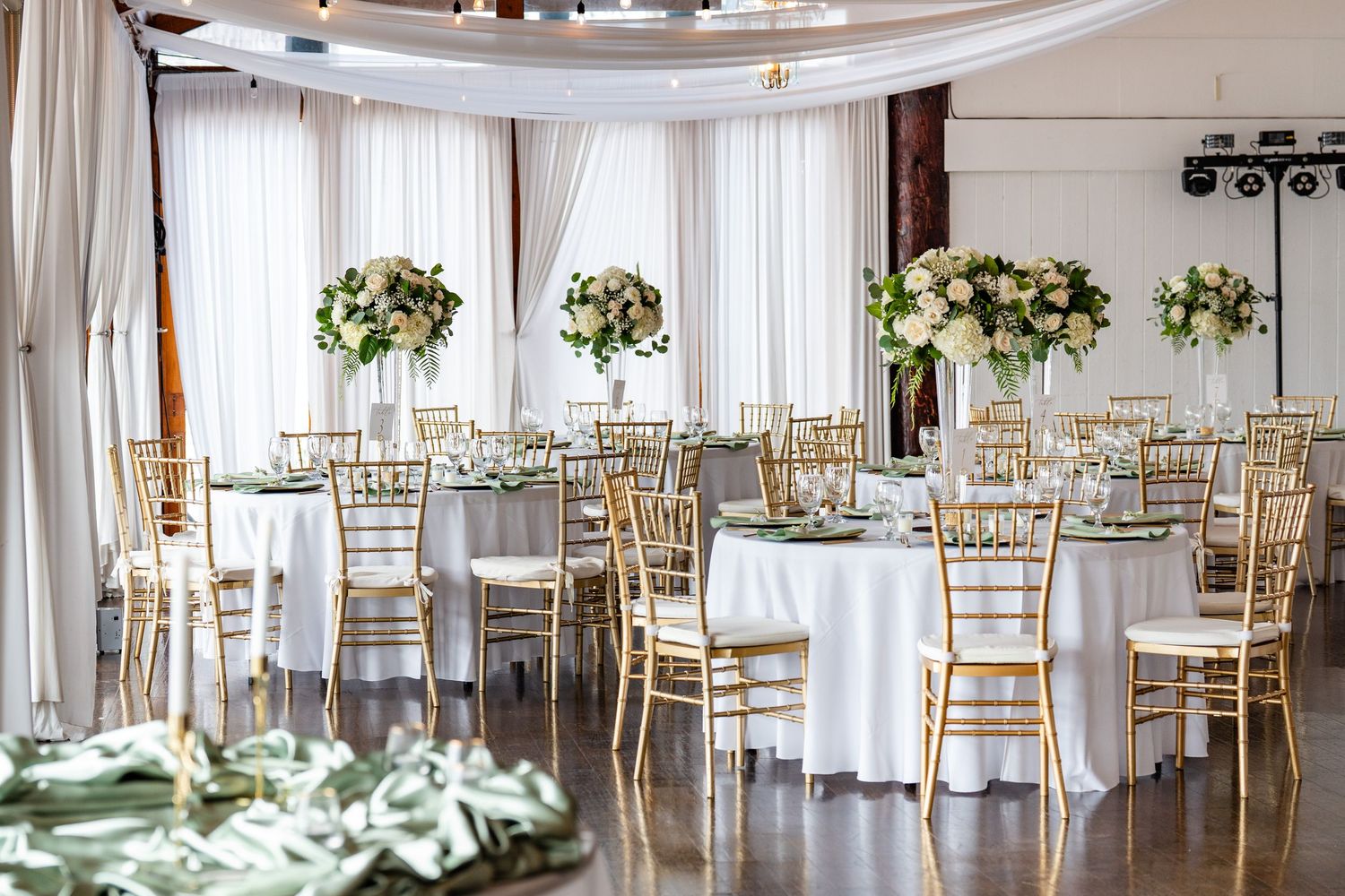 Elegant wedding reception hall with gold chiavari chairs, white tablecloths, and tall floral centerpieces in a draped venue.