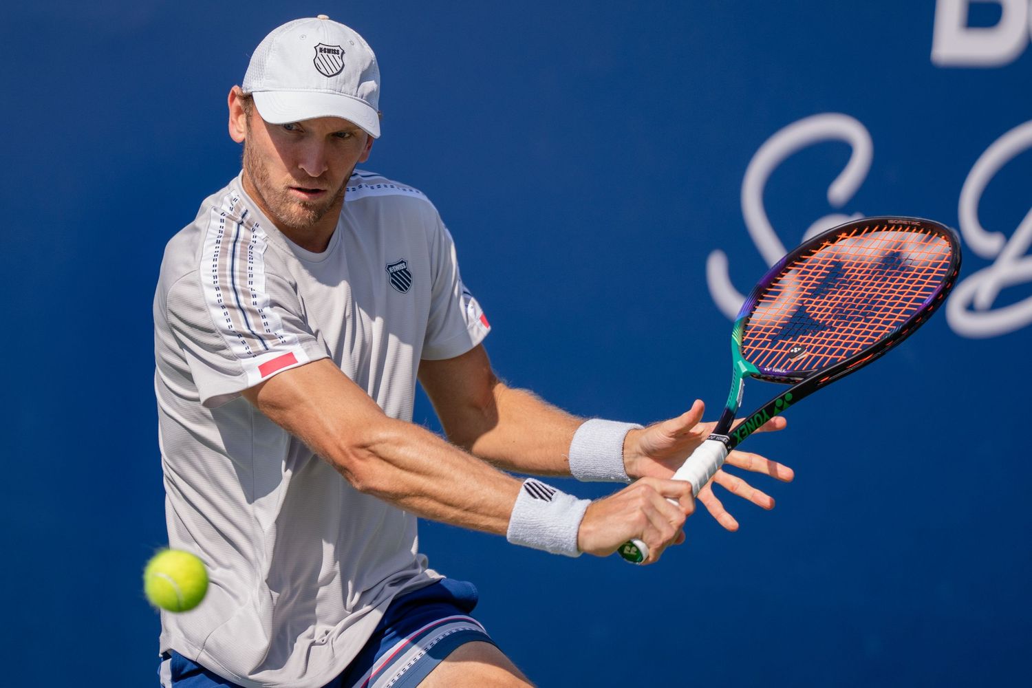Male tennis player in white hits a backhand shot against a blue background on hard court.