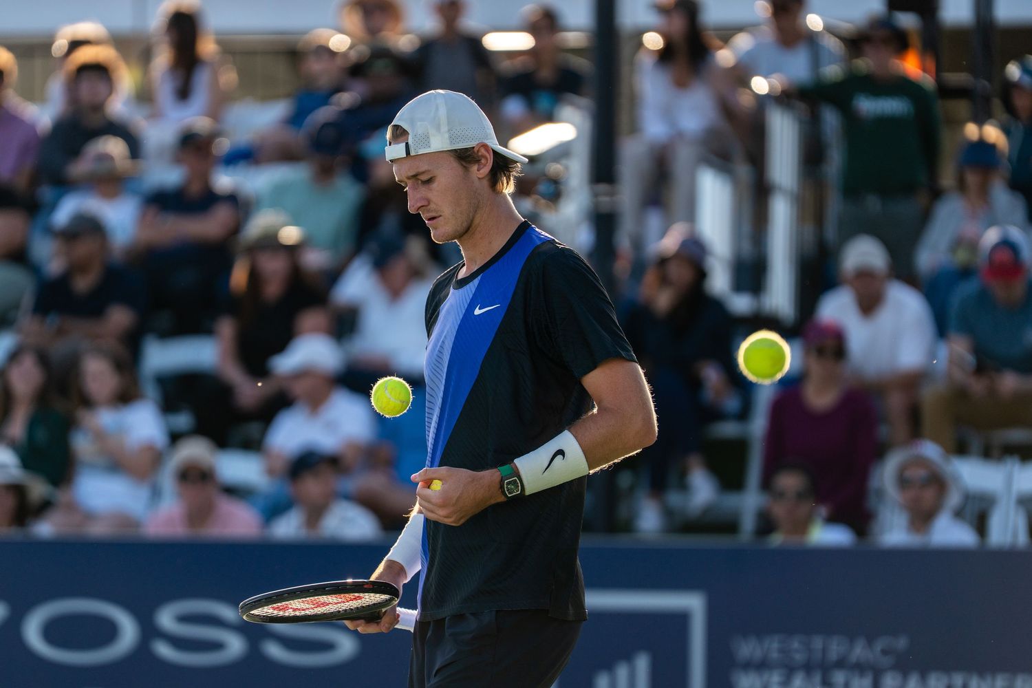 Male tennis player juggling two yellow balls while holding a racket during an outdoor tennis match.