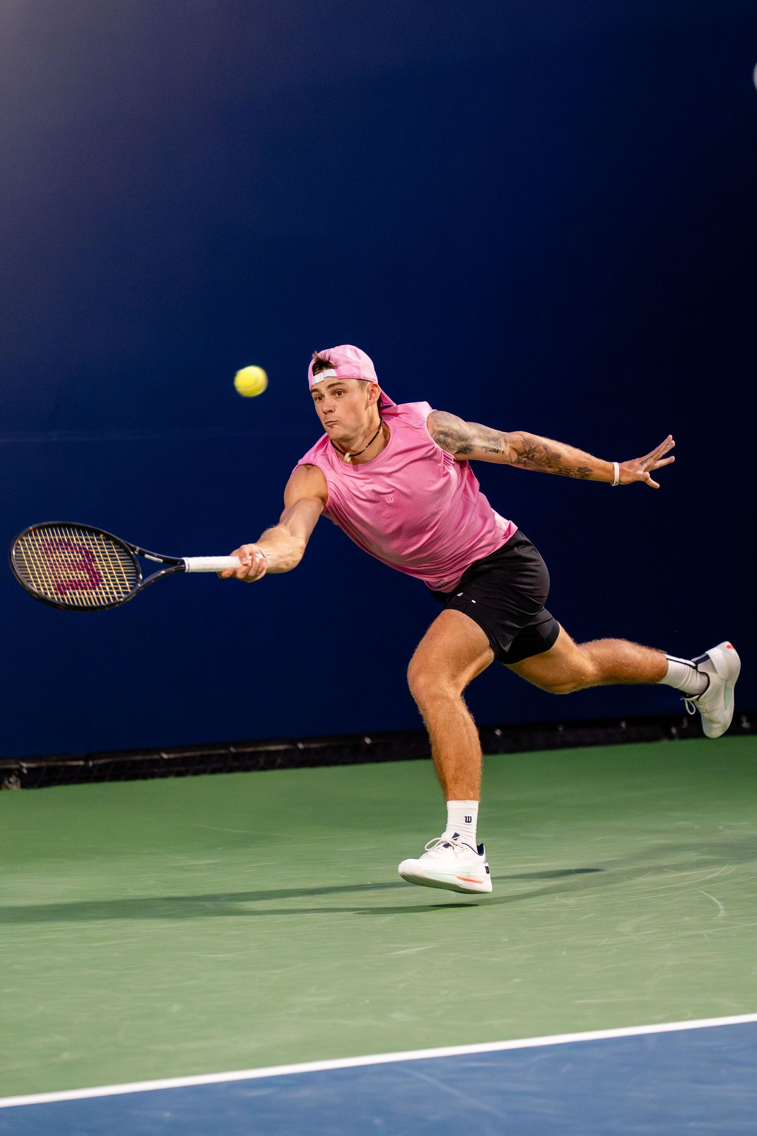Tattooed tennis player in pink tank top sprints to return a forehand shot on indoor blue court.