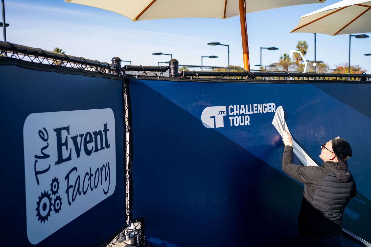 Worker installing ATP Challenger Tour branding on a navy blue court barrier at a tennis tournament.