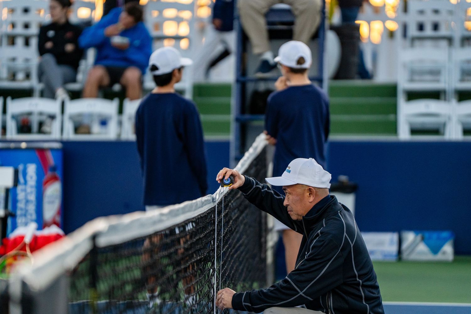 Official leans on the tennis net during an evening match at a professional outdoor tennis tournament.