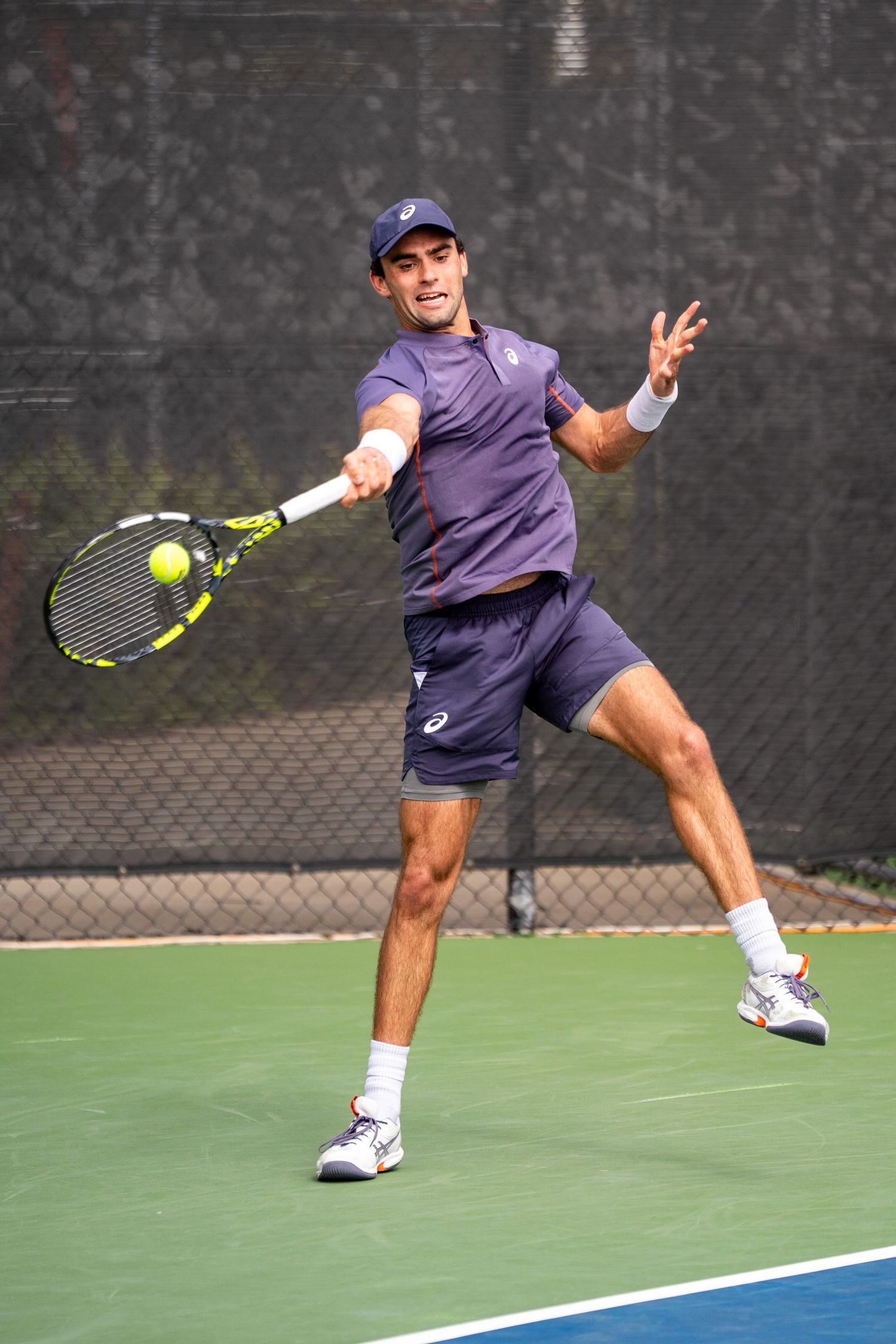 Tennis player in navy Asics outfit hits forehand shot on outdoor court.