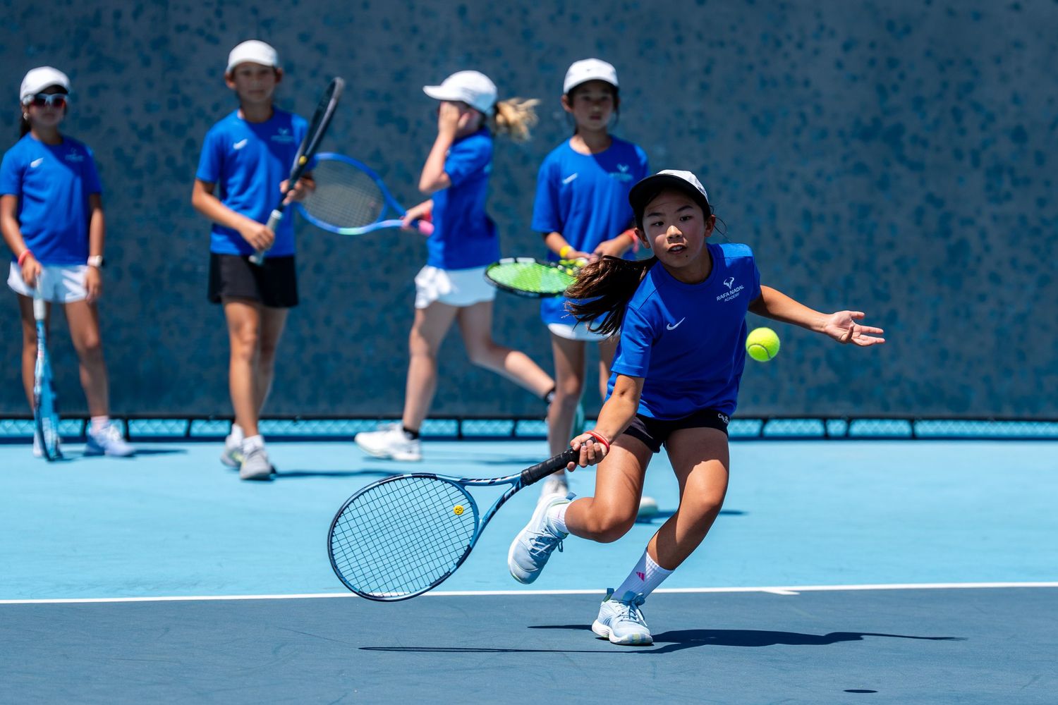 Young tennis players in blue uniforms practice on a bright court, with a player lunging to hit a forehand shot.