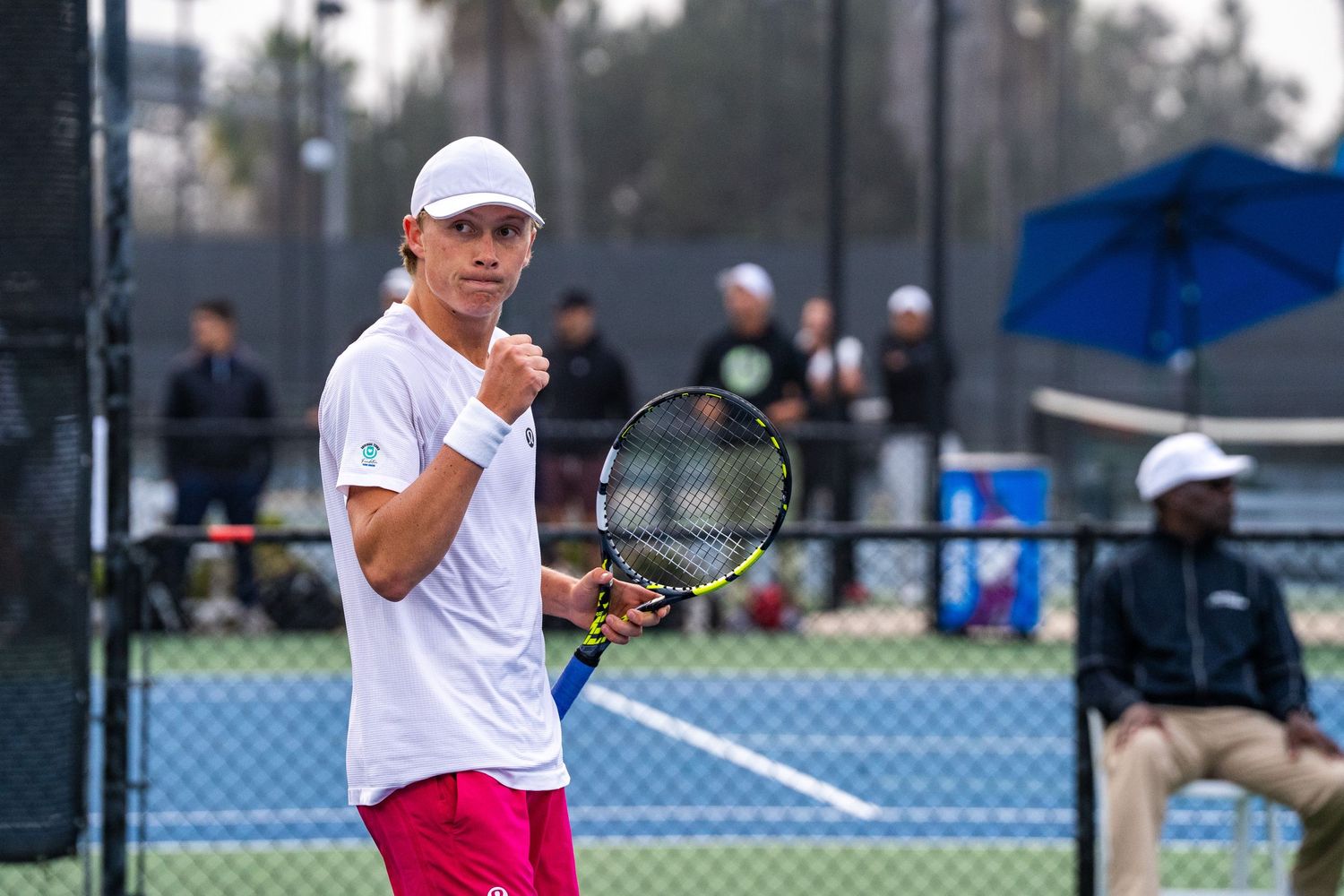 Young male tennis player celebrates a point wearing pink shorts on an outdoor court.