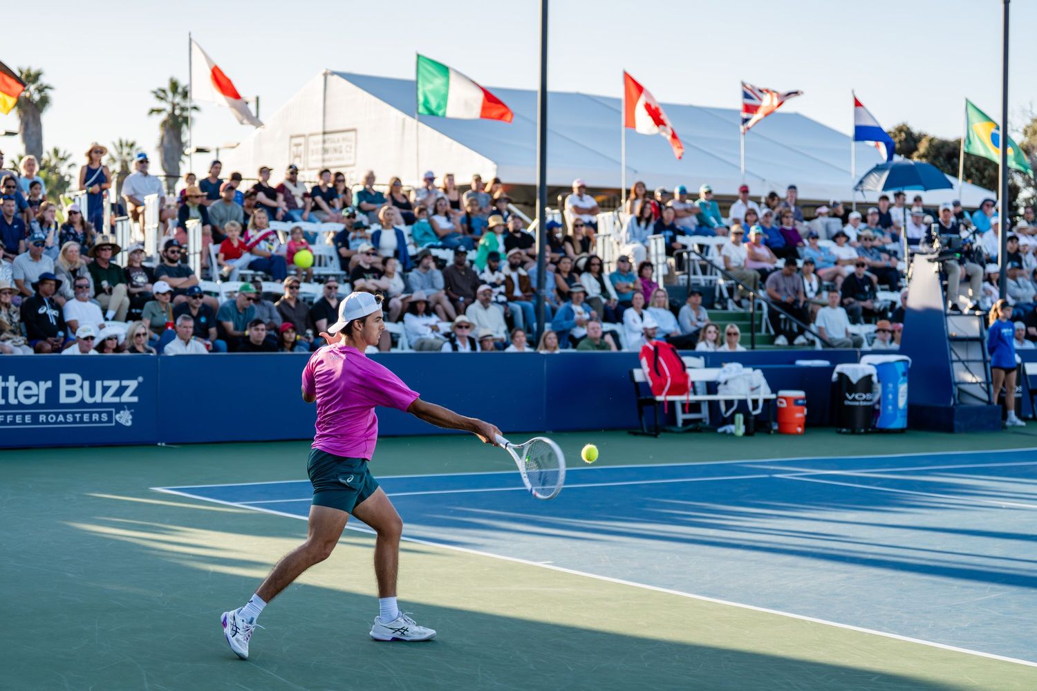 Tennis player in pink serves before a packed crowd with international flags at the San Diego Open.