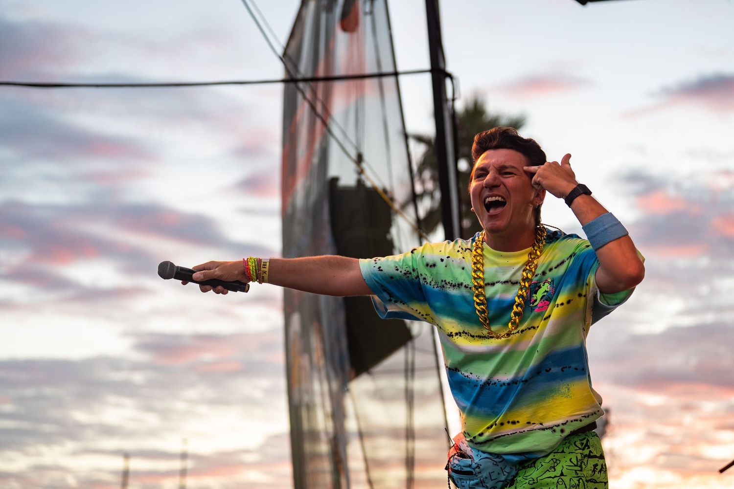 Performer in tie-dye shirt and gold chains entertains crowd at sunset outdoor festival.