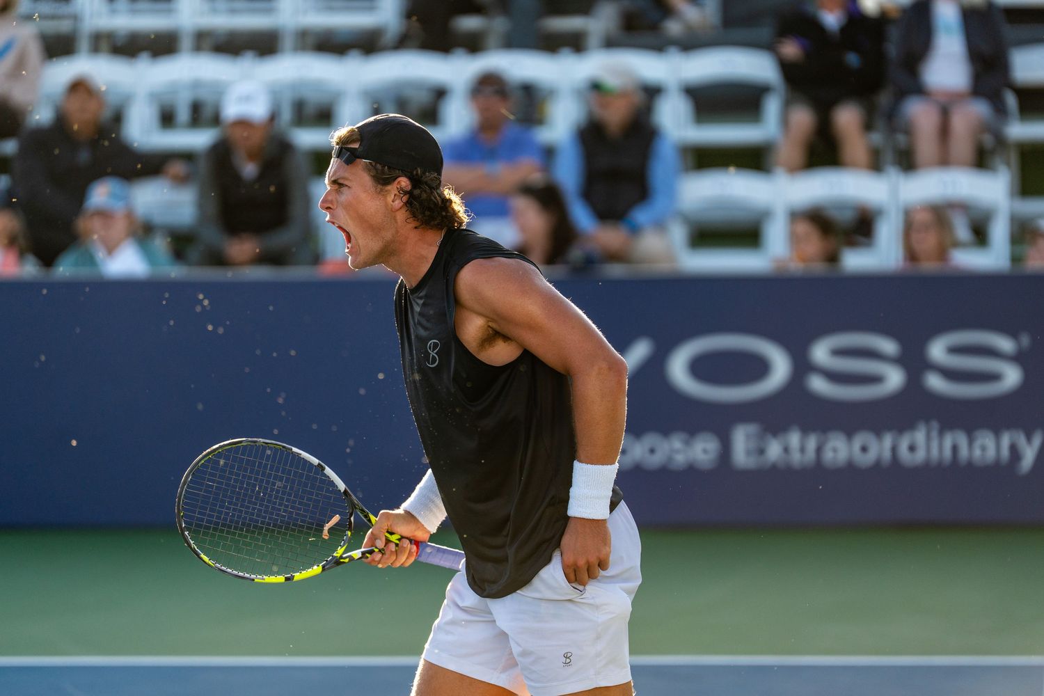 Tennis player celebrating with fist pump on court, wearing black outfit, with crowd visible in background.