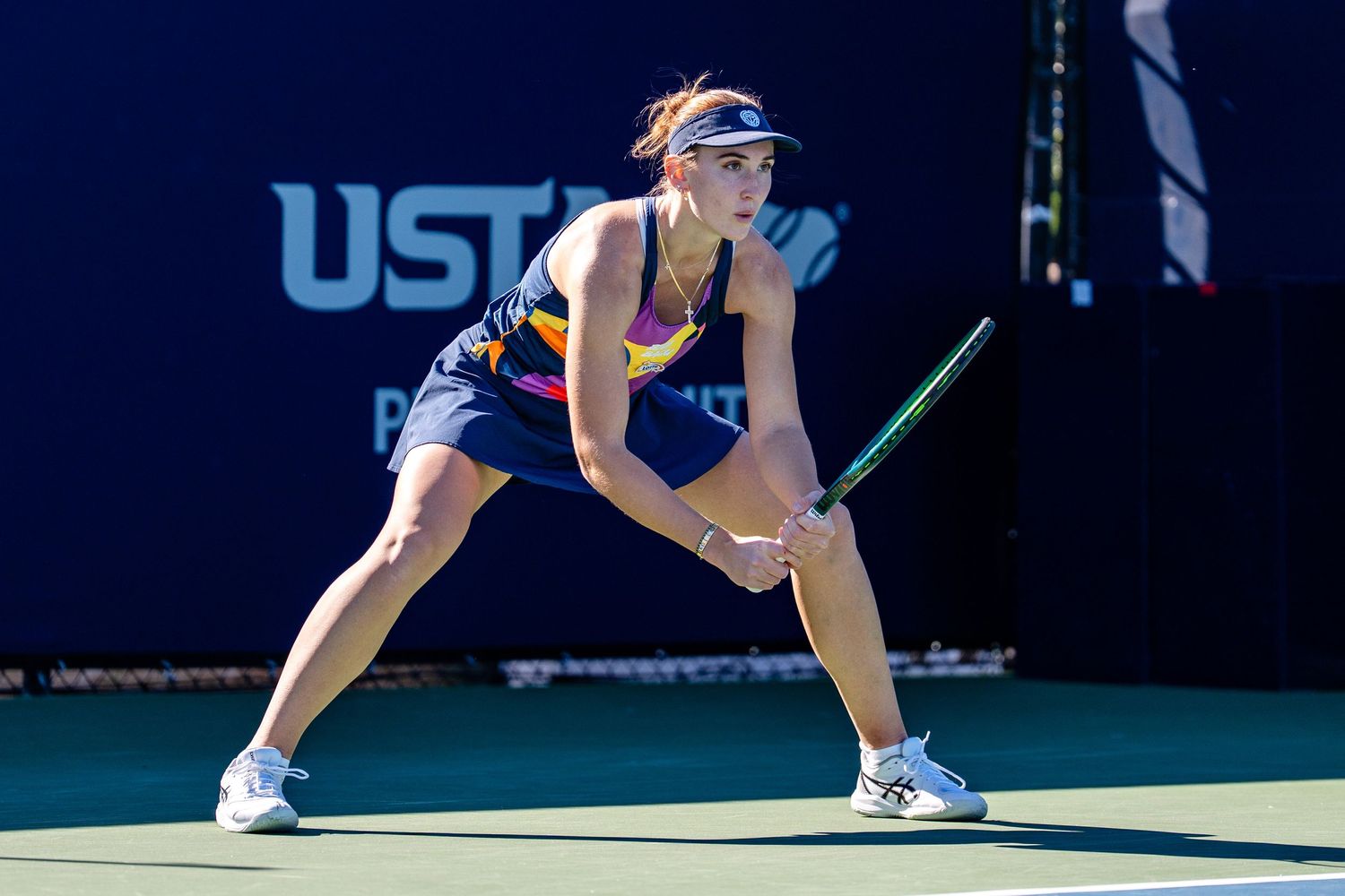 Female tennis player in navy outfit crouches in ready position on a USTA tennis court.