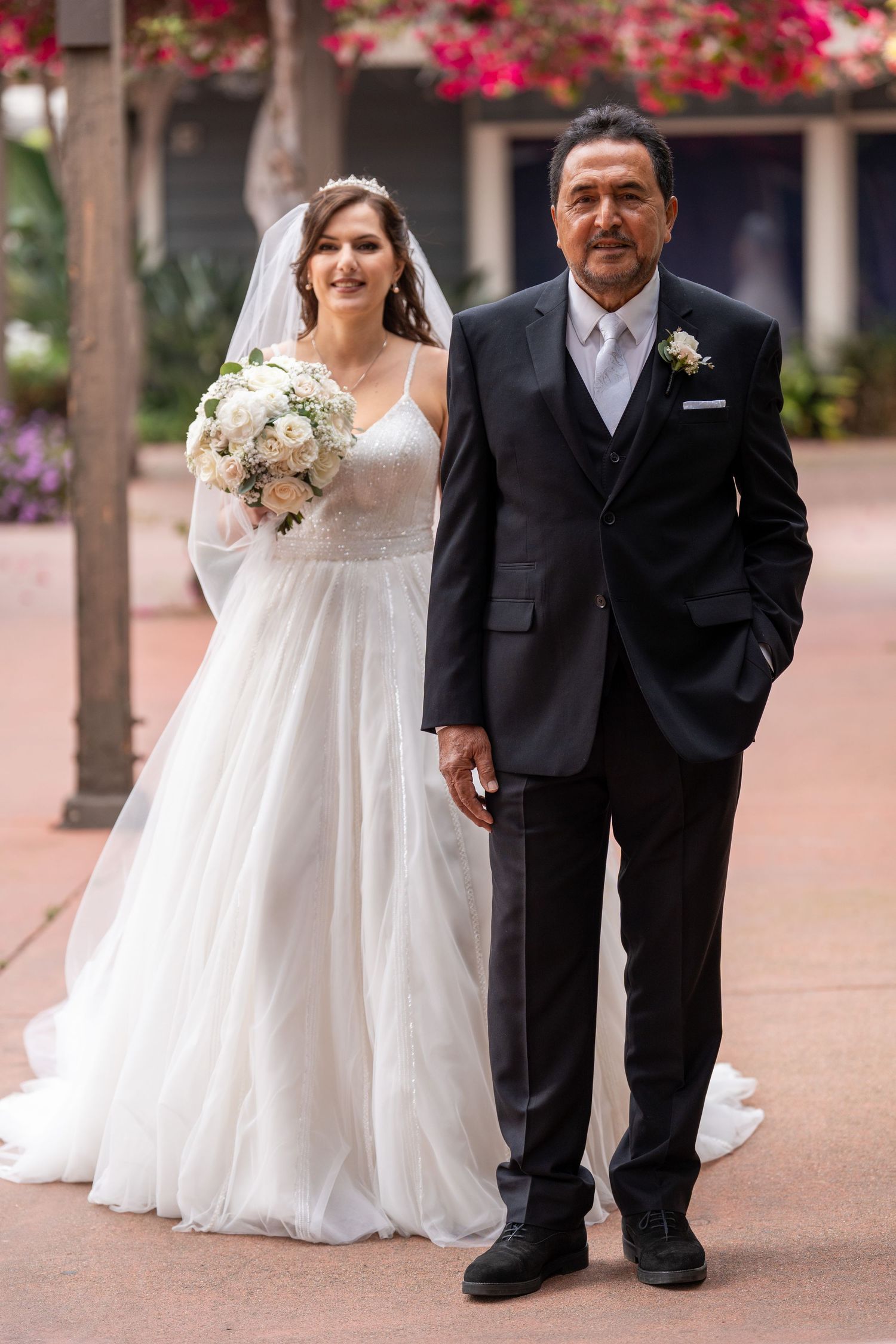 Bride in white gown holding bouquet walks with man in black suit at outdoor wedding venue with flowers.