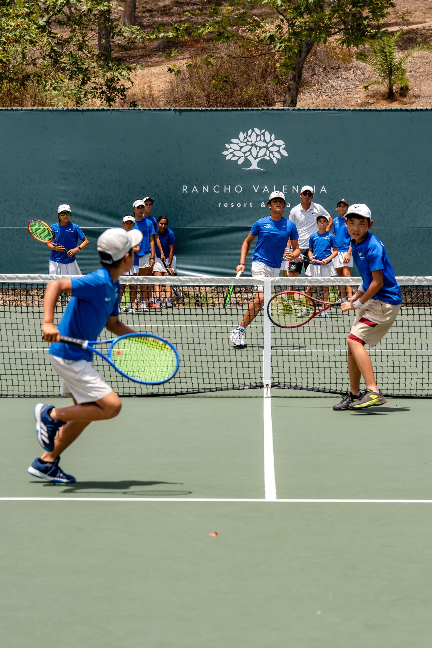 Young tennis players in blue uniforms practice on a court at Rancho Valencia Resort on a sunny day.