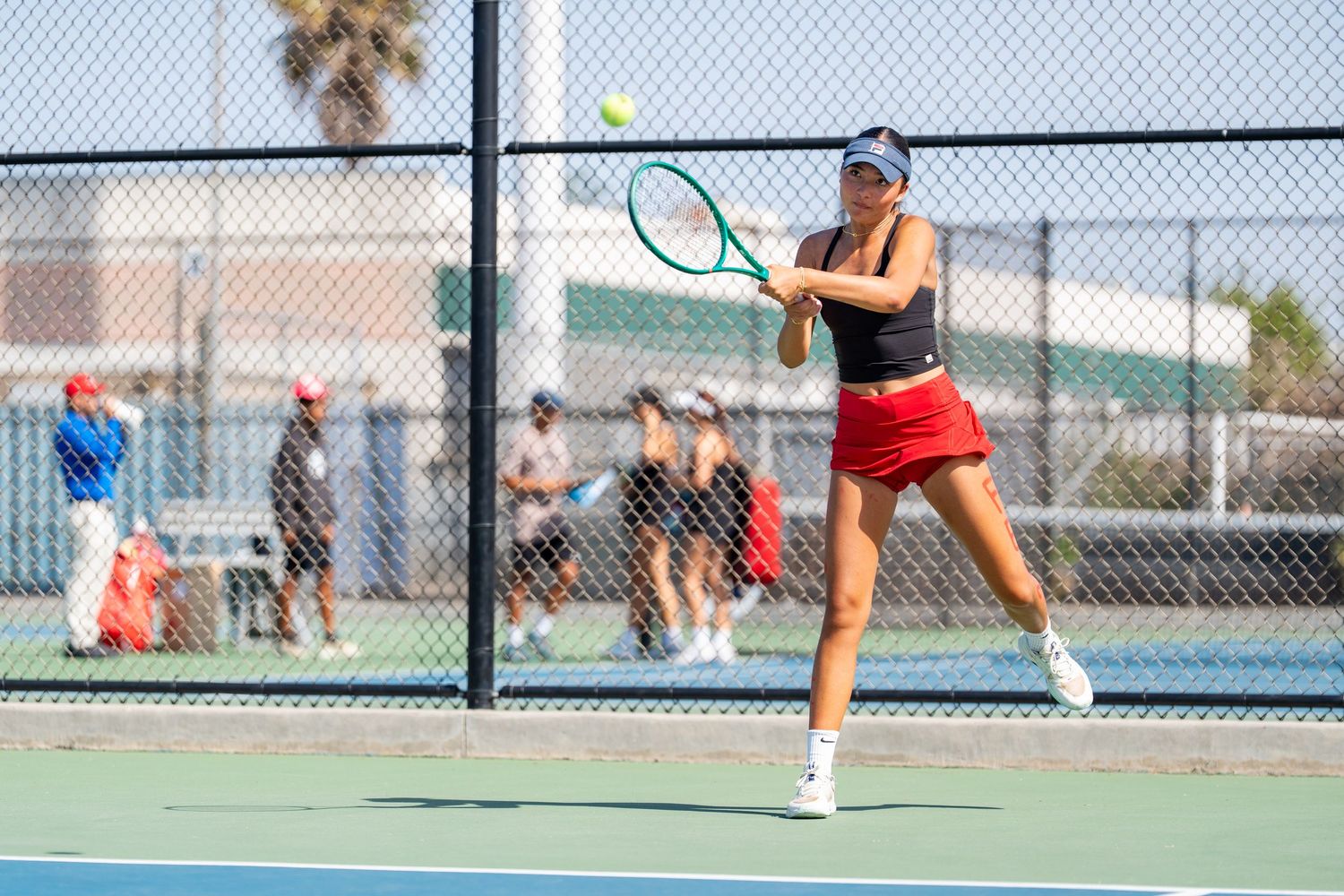 Female tennis player in black top and red skirt hits a forehand on an outdoor court with spectators behind.