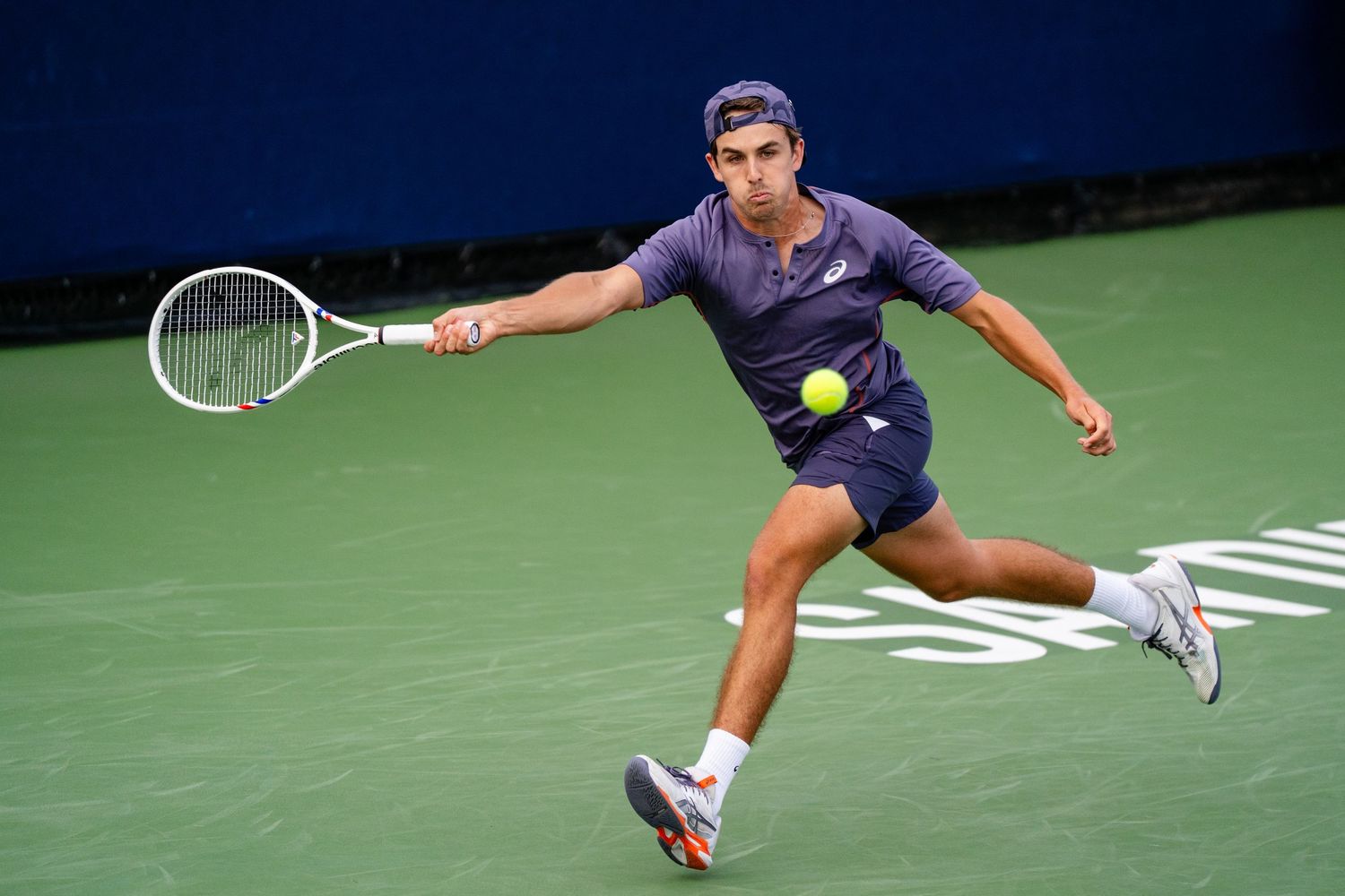 Tennis player in dark outfit lunges forward to return a shot on an outdoor green court.