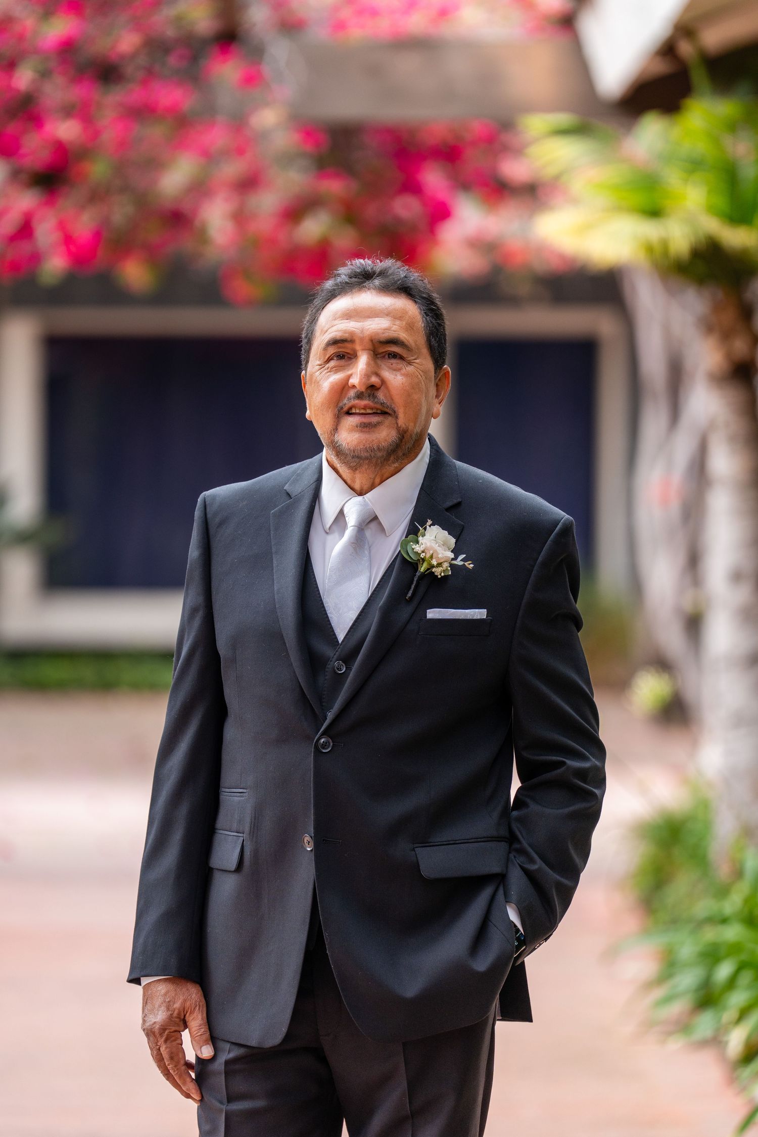 Elegant man in dark suit with boutonniere poses outdoors at wedding venue with pink flowers in background.