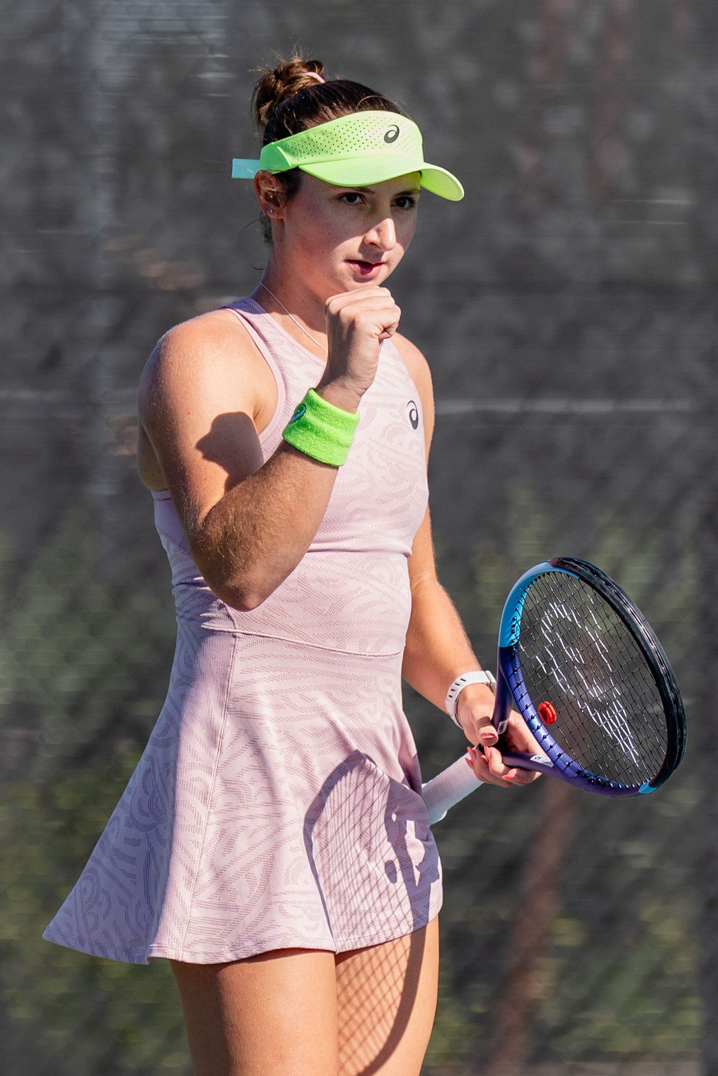 Female tennis player in pink dress holding racket wearing green ASICS visor during outdoor match.