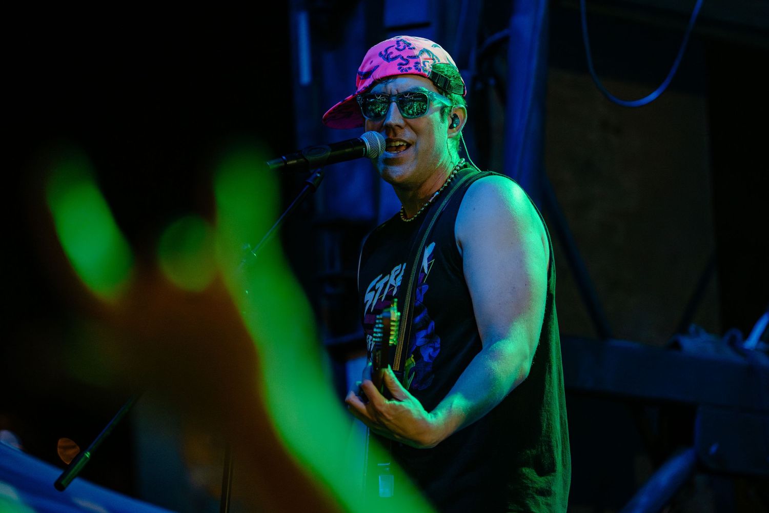 Male musician performs at microphone under colorful green stage lights at night concert.