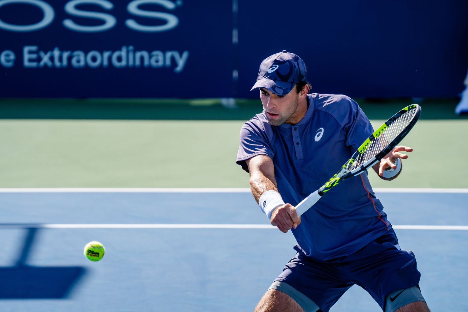 Male tennis player in blue ASICS kit hitting a backhand shot during an outdoor professional tennis match.