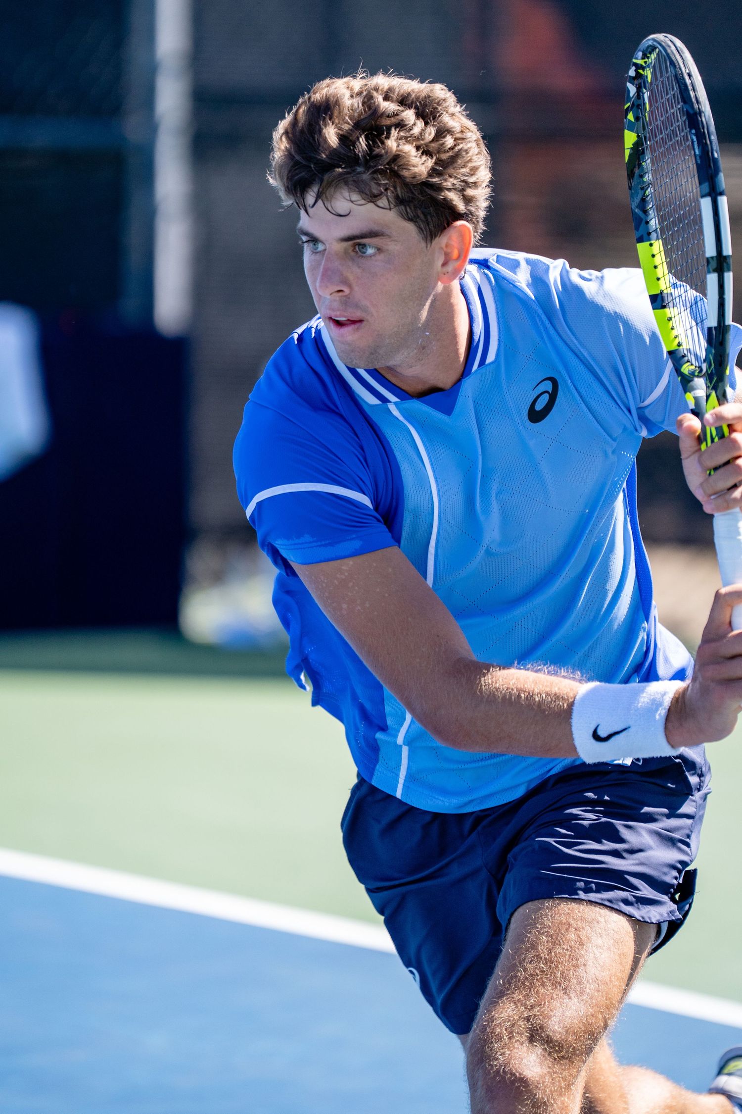 Male tennis player in blue ASICS shirt prepares to hit a backhand shot on an outdoor hard court.