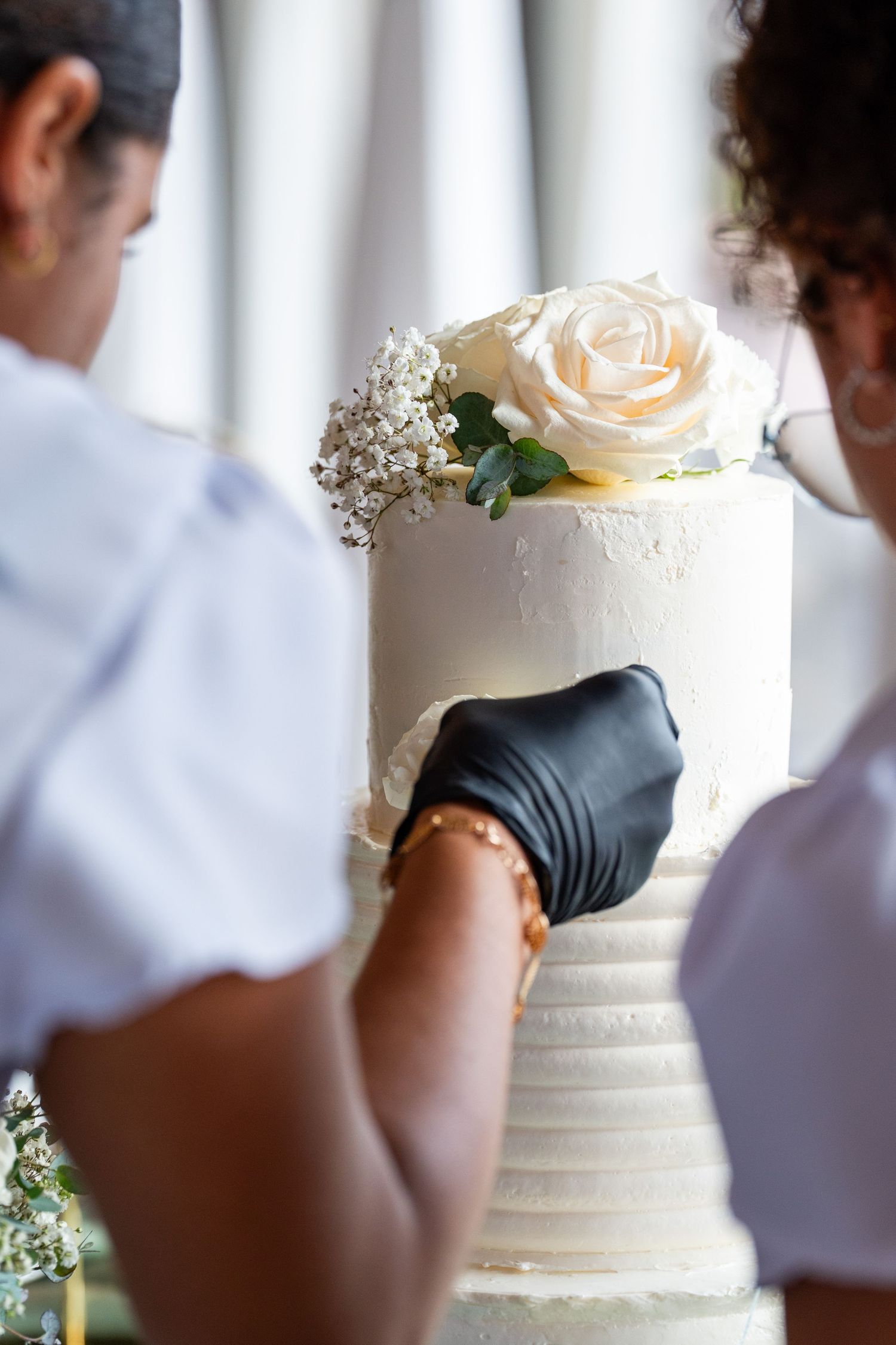 Two bakers in black gloves decorating a white wedding cake topped with a cream rose and baby's breath.
