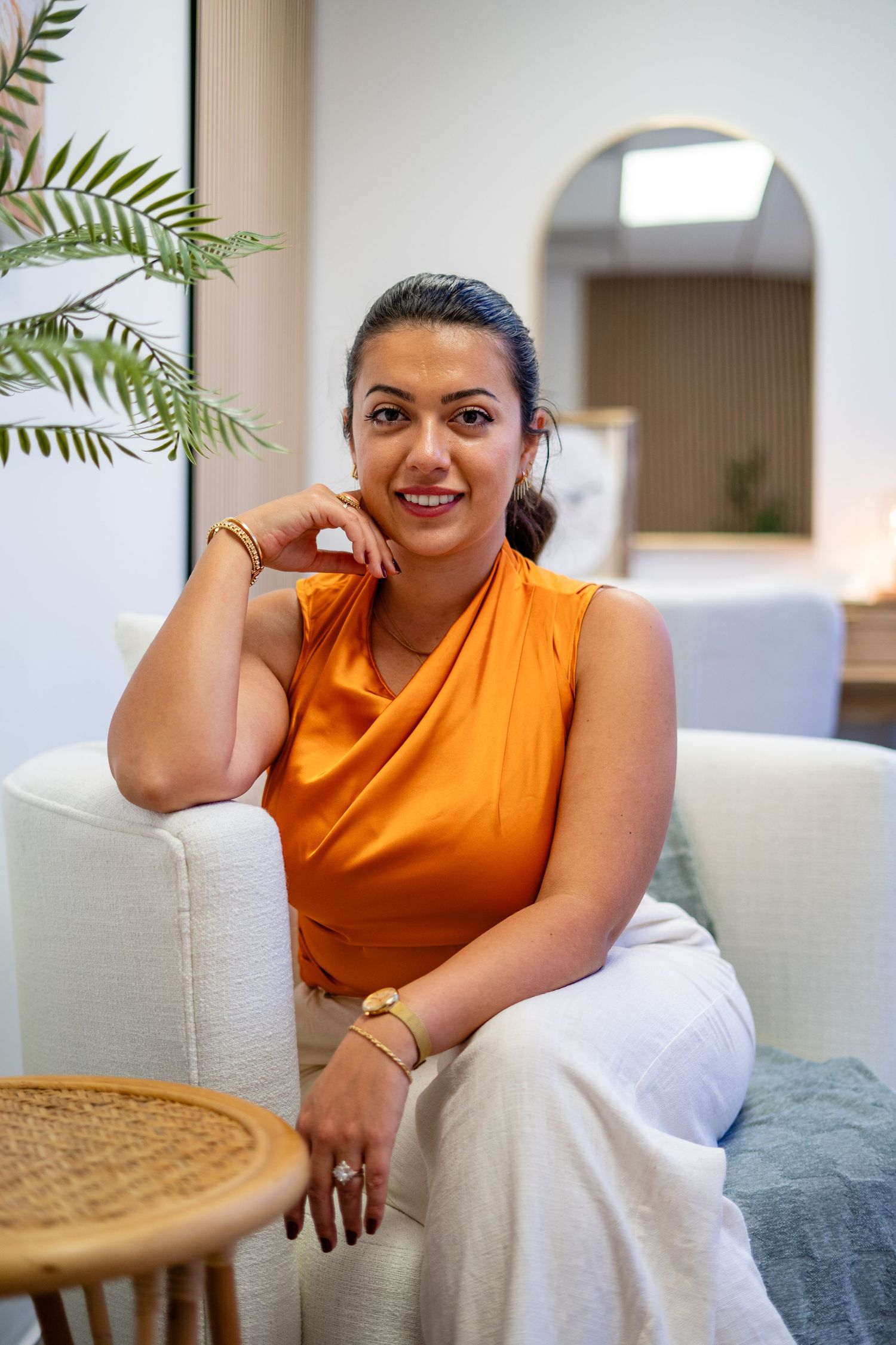Smiling woman in orange top and white pants posing in stylish modern room with arched mirror.