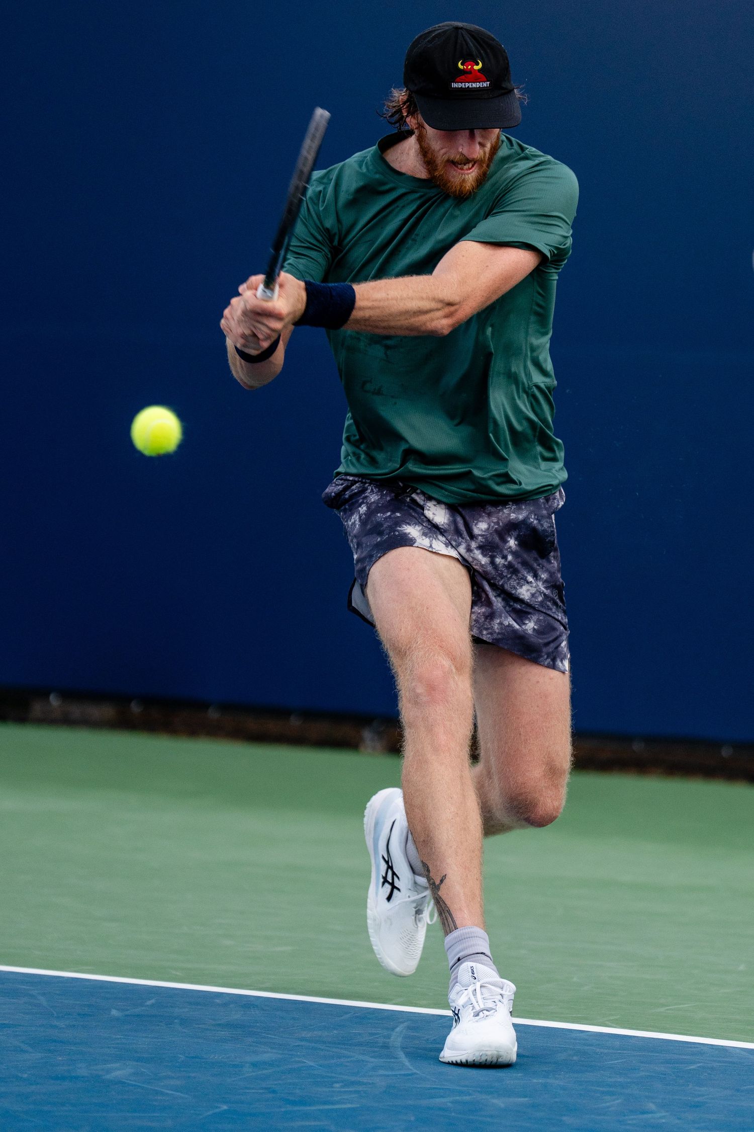 Male tennis player in green shirt and cap executing a powerful backhand stroke on a hard court.